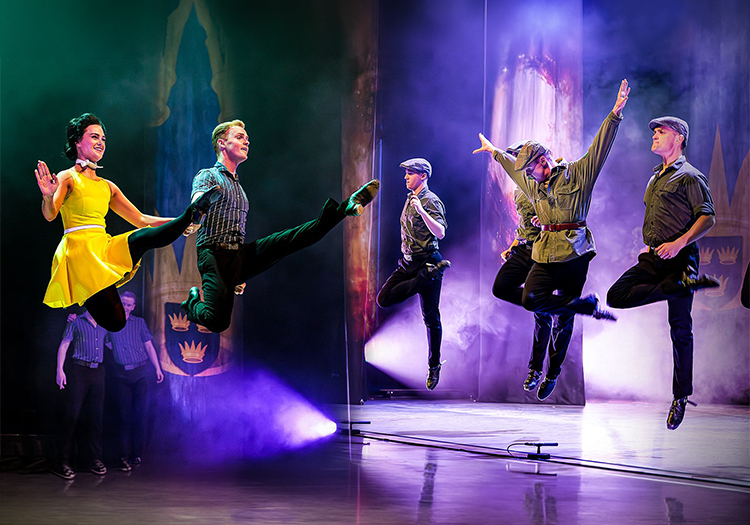 Dancers midair in a stage performance, with one in a yellow dress and others in caps, lit by colorful spotlights and haze.